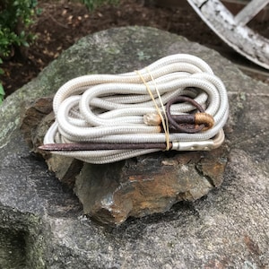 Könnte beinhalten: Ein weißes Seil mit einem Metallring und einem Metallhaken, zusammengebunden mit einem Gummiband. Das Seil ist aufgerollt und liegt auf einem grauen Stein.