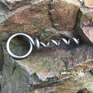 May include: A silver metal corkscrew with a ring at the top. The corkscrew is lying on a rough, brown rock.
