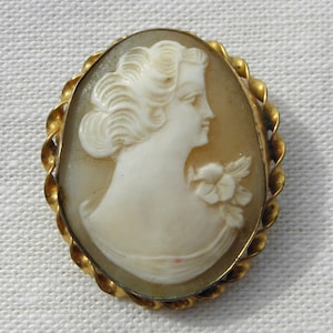 Pode incluir: Um broche de camafeu dourado com um camafeo branco de um perfil de mulher com uma flor no cabelo. O camafeu está engastado em uma moldura de design de corda torcida.