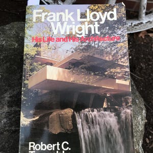 Op de afbeelding: Boekomslag van "Frank Lloyd Wright: His Life and His Architecture" door Robert C. Twombly. De afbeelding toont een gebouw met een waterval en bomen. De titel is in wit en rood.