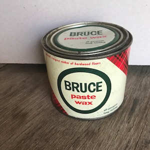 Op de afbeelding: Een vintage blik Bruce-pastawas. Het cilindrische blik heeft een witte achtergrond met een rood-zwart tartanpatroon. De tekst "BRUCE paste wax" is gedrukt in een groene cirkel. Het blik staat op een houten oppervlak.