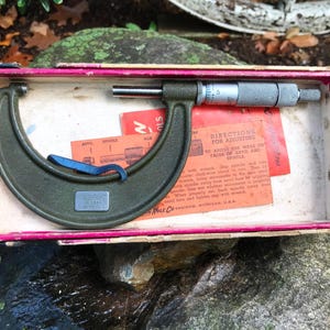 Puede incluir: Un micrómetro verde vintage con una escala de medición plateada, dentro de una caja forrada en rosa. La herramienta tiene un marco curvo y un husillo para mediciones precisas. Las instrucciones son visibles.