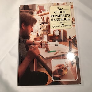 May include: The Clock Repairer's Handbook by Laurie Penman. The book cover shows a man repairing clocks, with a close-up inset of hands working on a clock. The cover has a beige background with the title in a cream-coloured box.