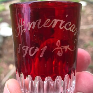 Op de afbeelding: Een rood glazen shotglas met een heldere bodem. Het glas is gegraveerd met de woorden "American 1901 25".