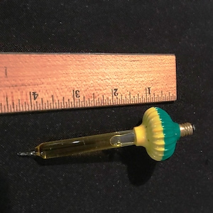 Może przedstawiać: Żarówka choinkowa ze szkła w kolorze żółtym i zielonym, z ostrym końcem i zaokrąglonym, wypukłym wierzchołkiem. Żarówka ma około 7,5 cm długości.