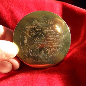 May include: A gold-coloured metal compass with a map of Florida and the word "Florida" in a banner across the top. The compass features the names of cities and towns in Florida, including Pensacola, Panama City, Jacksonville, Tallahassee, St. Augustine, Silver Spring, Marine Land, Orlando, Daytona Beach, St. Petersburg, Sarasota, Clearwater, Palm Beach, Cypress Gardens, Hollywood, Miami Beach, Bok Tower, and Key West.
