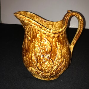 Pode incluir: Um jarro de cerâmica marrom e amarelo com um design floral. O jarro tem uma boca larga e uma alça.