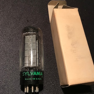 Puede incluir: Un tubo de vacío vintage con una base negra y un tubo de vidrio transparente. El tubo tiene una etiqueta verde que dice "SYLVANIA" y "MADE IN U.S.A.". El tubo está en su caja original.
