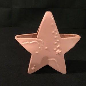 Puede incluir: Un macetero de cerámica en forma de estrella rosa con un diseño en relieve de estrellas y nubes. El macetero tiene un asa en la parte superior.