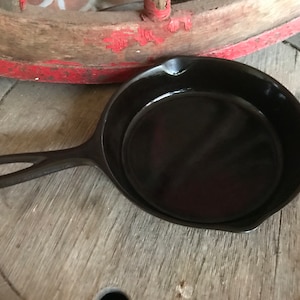 Peut inclure: Une poêle en fonte noire avec une courte poignée. La poêle est posée sur une surface en bois.