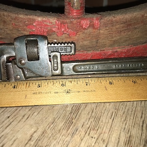 May include: An 8-inch, silver-colored pipe wrench with the text "TRIMCO DROP FORGED" and "8 TRIMCO" on the handle. The wrench is measured against a wooden ruler.