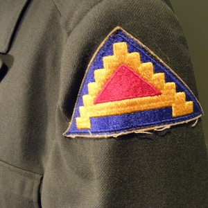 May include: A military uniform patch with a blue, yellow, and red triangle design. The patch is sewn onto a dark green fabric.
