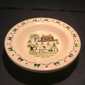 Puede incluir: Cuenco de cerámica color crema con una escena rural que representa una casa roja, un árbol y un granjero arando con caballos. El borde tiene un patrón de hojas y puntos verdes y rojos. Se ve una pila de cuencos.
