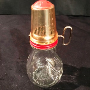 May include: A vintage glass shaker with a red and gold lid and a metal handle. The shaker has a textured glass body and a red plastic top with a clear plastic lid.