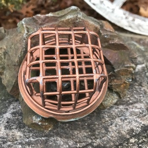 Op de afbeelding: Een koperkleurige metalen kooi met een ronde basis, die op een rots ligt. De kooi heeft een vierkante vorm en een gaasontwerp.