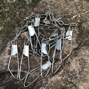 Puede incluir: Un montón de eslabones de cadena de metal plateado con un gancho en cada extremo. Los eslabones de la cadena están dispuestos en un patrón aleatorio.