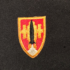 Puede incluir: Un parche bordado en rojo, blanco y dorado con un misil negro y una nube blanca. El parche tiene forma de escudo.