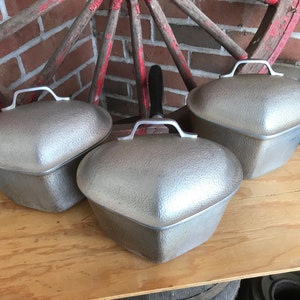 May include: Three silver heart-shaped aluminum casserole dishes with lids. The dishes have a textured surface and are arranged on a wooden surface.