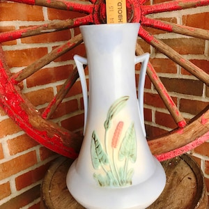 Peut inclure: Un vase en céramique bleu clair avec un motif floral. Le vase a deux anses et une base évasée. Le motif floral présente des feuilles vertes et des fleurs roses.
