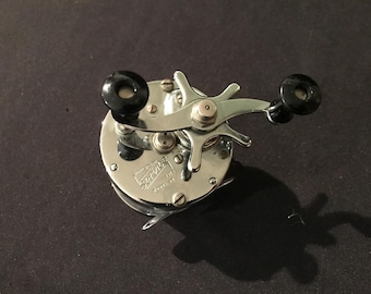 May include: A vintage silver fishing reel with black handles. The reel has a circular body with a handle and a star-shaped drag adjustment. The word "Service" is printed on the reel.