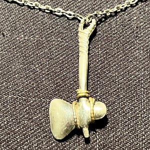 May include: A silver-toned metal pendant shaped like a small axe or tomahawk, suspended from a matching silver-toned chain. The axe head is secured to the handle with thin, coiled gold-toned wire, and the handle features a textured, wrapped design.