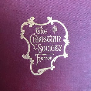 May include: Close-up of a deep purple book cover with gold lettering. The text reads "The Christian Society Herron" in an ornate, stylized font, framed by decorative flourishes. The cover has a subtle, textured pattern.