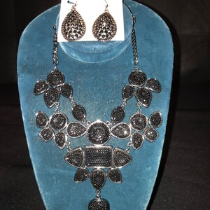 Peut inclure: Un ensemble collier et boucles d'oreilles en strass noirs, présenté sur un buste en velours bleu. Le collier présente un grand motif central avec des formes géométriques et un pendentif en forme de goutte. Les boucles d'oreilles sont en forme de goutte.