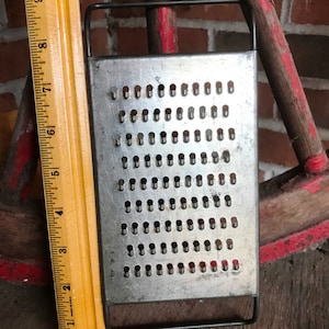 May include: A vintage metal grater with a black handle. The grater has a rectangular shape and is made of metal with small holes. The grater is about 6 inches long and 3 inches wide.