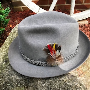 Op de afbeelding: Een grijze fedora hoed met een gevlochten band en een klein veren accent. De veer is rood, geel en bruin.