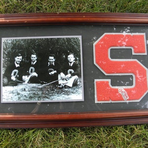 May include: Framed vintage photograph of a group of men in athletic uniforms, with a large orange and white letter "S" emblem. The photograph is in a dark brown wooden frame, resting on green grass.