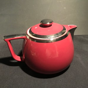 May include: A maroon ceramic teapot with a silver and black trim around the lid. The teapot has a round body and a black handle.