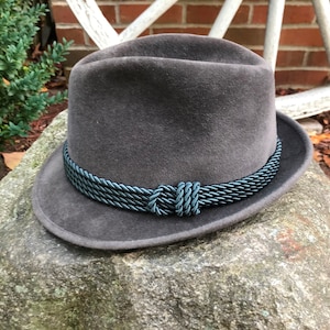 Puede incluir: Un sombrero fedora de fieltro gris con una banda trenzada azul oscuro alrededor de la corona.