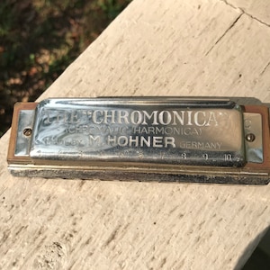 Puede incluir: Una armónica cromática Hohner 'The Chromonica' de color plateado con el texto 'Made by M. Hohner Germany' en la parte delantera.