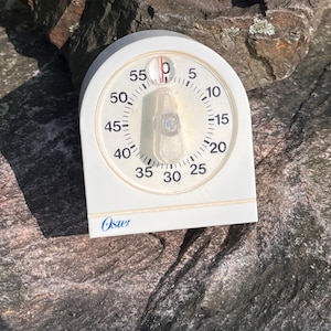 May include: An off-white Oster kitchen timer with a rounded top and a circular analog dial featuring black numbers from 0 to 55. A central clear plastic knob sits over the dial, and the brand name Oster is printed in blue on the lower front edge.