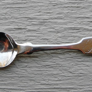 May include: A silver teaspoon with a rounded bowl and a long, slender handle. The handle is engraved with the initials "B.C.J."