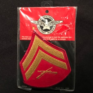 以下が含まれることがあります： 赤と金の軍用章のワッペンで、金色の2本の山形と交差したライフルが描かれています。ワッペンは、赤と白のラベルが付いたプラスチック製の袋に入っています。ラベルには「メーカー Hilborn-Hamburger Inc. 軍用章」と書いてあります。