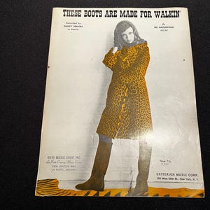 Op de afbeelding: Een vintage bladmuziekomslag voor These Boots Are Made For Walkin' met een persoon in een jas met luipaardprint en bruine kniehoge laarzen. De tekst bevat credits voor Nancy Sinatra en Lee Hazlewood, plus details van Roxy Music Shop.