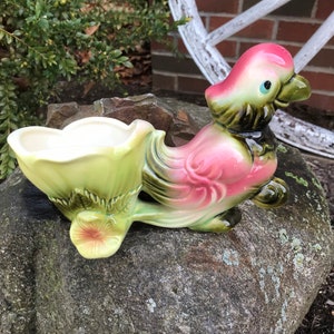 Może przedstawiać: Wazon ceramiczny w stylu vintage w kształcie różowo-zielonego ptaka z doniczką na grzbiecie. Wazon stoi na kamieniu.