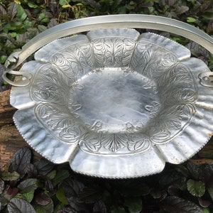 Op de afbeelding: Een zilveren aluminium serveerschaal met een decoratief bloemmotief en een draadhandvat. De schaal staat op een houten oppervlak.