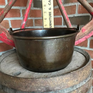 May include: A vintage cast iron pot with a wire handle. The pot is dark brown and has a worn, rustic look. It is sitting on a wooden barrel.