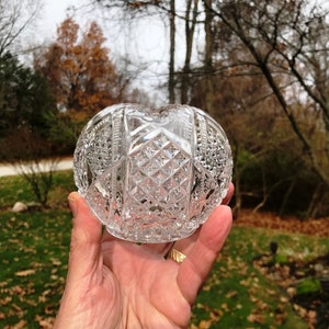 May include: A clear glass bowl with a cut glass design. The bowl is round and has a wide opening. It is held in a hand with a gold ring.