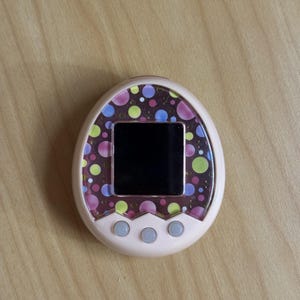 Könnte beinhalten: Ein hellrosa, eiförmiges digitales Haustiergerät mit einem dunklen Bildschirm, der von einem dekorativen Rand aus bunten Punkten in Lila, Blau, Gelb und Pink umgeben ist. Unter dem Bildschirm befinden sich drei kleine, runde, hellgraue Tasten.