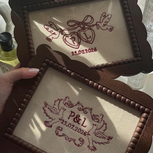 May include: Two rectangular wooden frames with beaded inner edges, each containing cream-coloured fabric with red embroidery. The top frame shows two birds holding hearts with the letters P and L and the date 25.07.2026. The bottom frame features two birds, the letters P&L, and the date 25.07.2026.