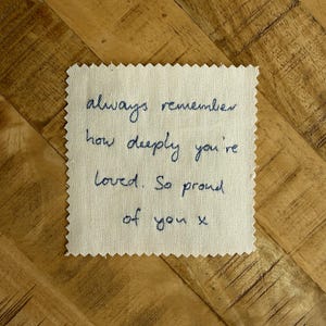May include: A square piece of off-white fabric with pinked edges featuring dark blue embroidered text that reads: always remember how deeply you're loved. So proud of you x. The embroidery is done in a cursive script style on the woven textile.