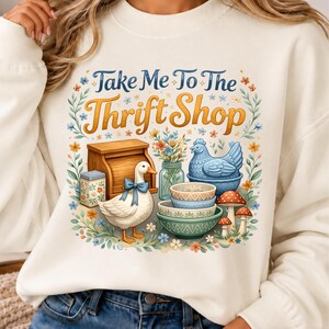Pode incluir: Uma camisola de gola redonda cor de creme com uma impressão gráfica com o texto Take Me To The Thrift Shop. A ilustração inclui um ganso branco com um laço azul, uma caixa de pão de madeira, uma estatueta de galinha azul, taças e cogumelos.
