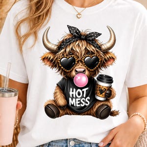 May include: A white t-shirt featuring a graphic of a brown Highland cow wearing a black polka-dot bandana and leopard-print heart-shaped sunglasses. The cow is blowing a pink bubble, wearing a black shirt with the text HOT MESS, and holding a cup.