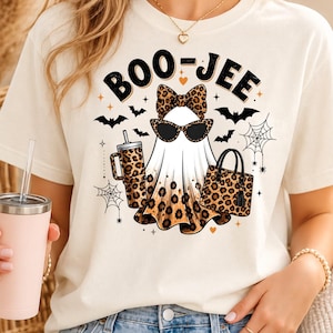 May include: A cream-colored t-shirt featuring a graphic of a ghost wearing a leopard print bow and sunglasses. The ghost holds a leopard print tumbler and handbag. The text reads BOO-JEE, surrounded by black bats, spiderwebs, and small hearts.
