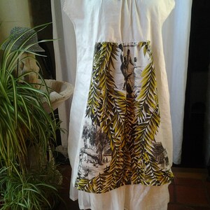 May include: A white linen dress with a gathered neckline and a printed panel featuring a tropical scene with palm trees and people. The dress has a white cotton slip underneath.