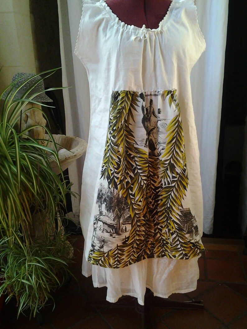 May include: A white linen dress with a gathered neckline and a printed panel featuring a tropical scene with palm trees and people. The dress has a white cotton slip underneath.