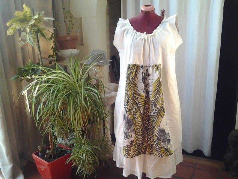 May include: A white linen dress with a floral print panel. The dress has a gathered neckline and a loose, flowing silhouette.